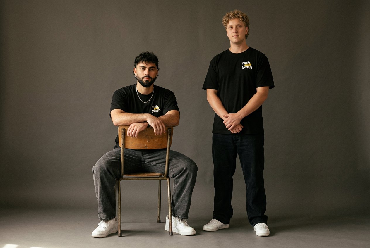 Bronson, seated on a wooden chair, and Tristan, standing, both in black Nah Yeah tees against a grey studio backdrop